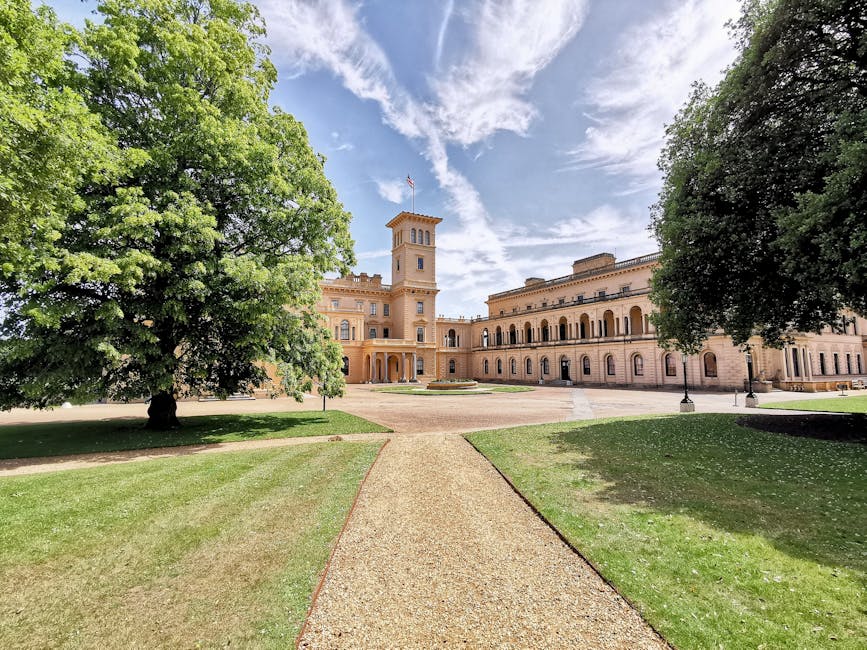 A wide, well-maintained gravel pathway runs directly toward a large, historic building with classical architecture, featuring a central tower topped with a flagpole. On either side of the pathway, the