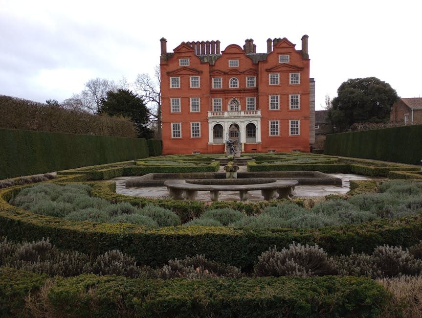 A wide, well-maintained gravel pathway runs directly toward a large, historic building with classical architecture, featuring a central tower topped with a flagpole. On either side of the pathway, the